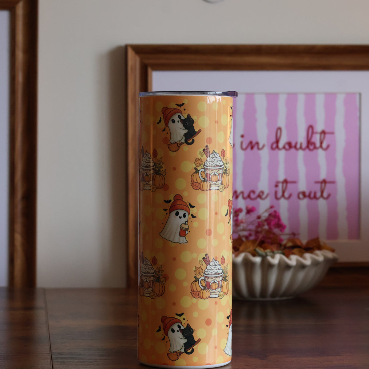 A tall cylindrical tin with a yellow and orange cupcake pattern stands on a wooden table in front of framed wall art.