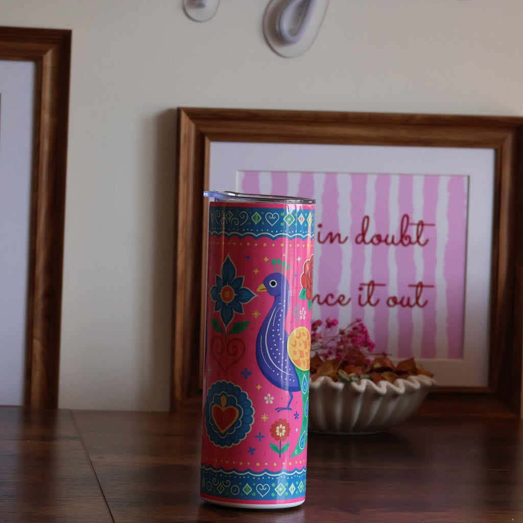 A colorful cylindrical tin with a peacock and floral pattern stands on a wooden table in a styled room setting, flanked by two framed art prints on a beige wall.
