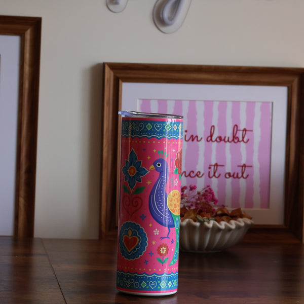 A colorful cylindrical tin with a peacock and floral pattern stands on a wooden table in a styled room setting, flanked by two framed art prints on a beige wall.