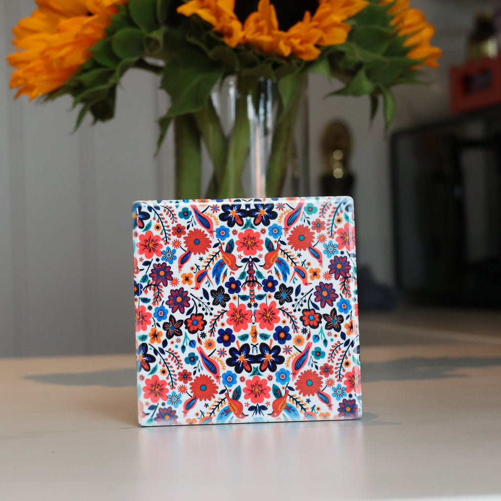 A square ceramic tile coaster with a colorful floral and geometric pattern, standing upright on a light-colored tabletop with a blurred vase of sunflowers in the background.