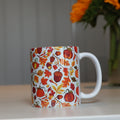 A white ceramic mug decorated with a colorful autumn pattern of pumpkins, acorns, and leaves, sitting on a light-colored tabletop with a blurred vase of orange flowers in the background.